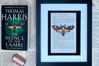 A framed art print featuring a printed moth on a page from Thomas Harris's book 'The Silence of the Lambs' is displayed next to the paperback book and a spool of pink thread on a background of vintage book pages.